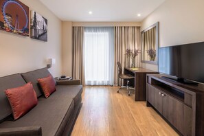 A modern hotel room with a sofa, a desk, a television, and a large window with curtains.