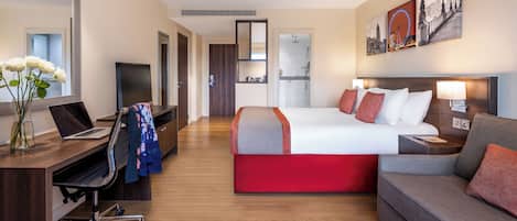 A hotel room with a bed, a desk, a chair, a sofa, a TV, and artwork on the walls.