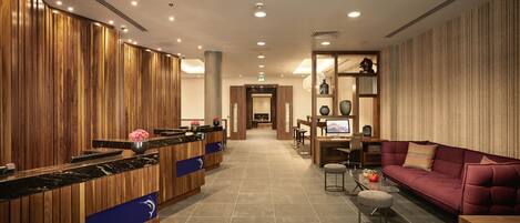 A modern hotel lobby with a wooden reception desk, a seating area with a sofa and armchairs, and a dining area with a table and chairs.