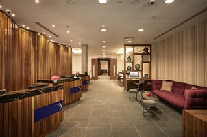 Eine moderne Hotelhalle mit einem Holzbuchtschalter, einer Sitzecke mit Sofa und Sesseln sowie einem Essbereich mit Tisch und Stühlen.