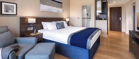 A modern hotel room with a large bed, a seating area, a desk, and a bathroom visible through an open door.