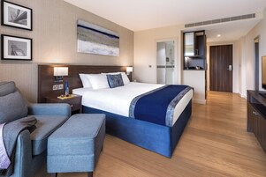 A modern hotel room with a large bed, a seating area, a desk, and a bathroom visible through an open door.