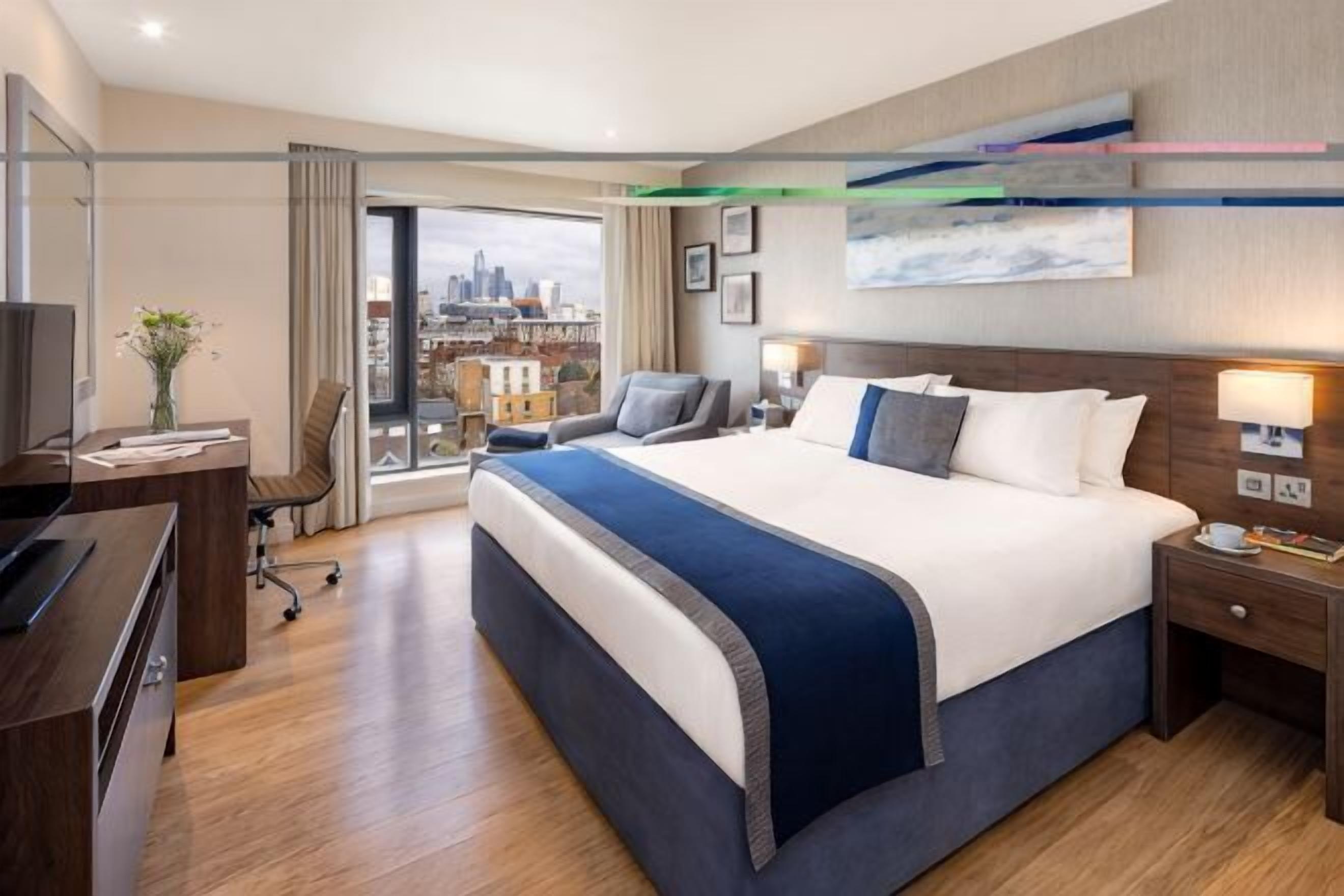 A hotel room with a large bed, a desk, a chair, a TV, and a city view.
