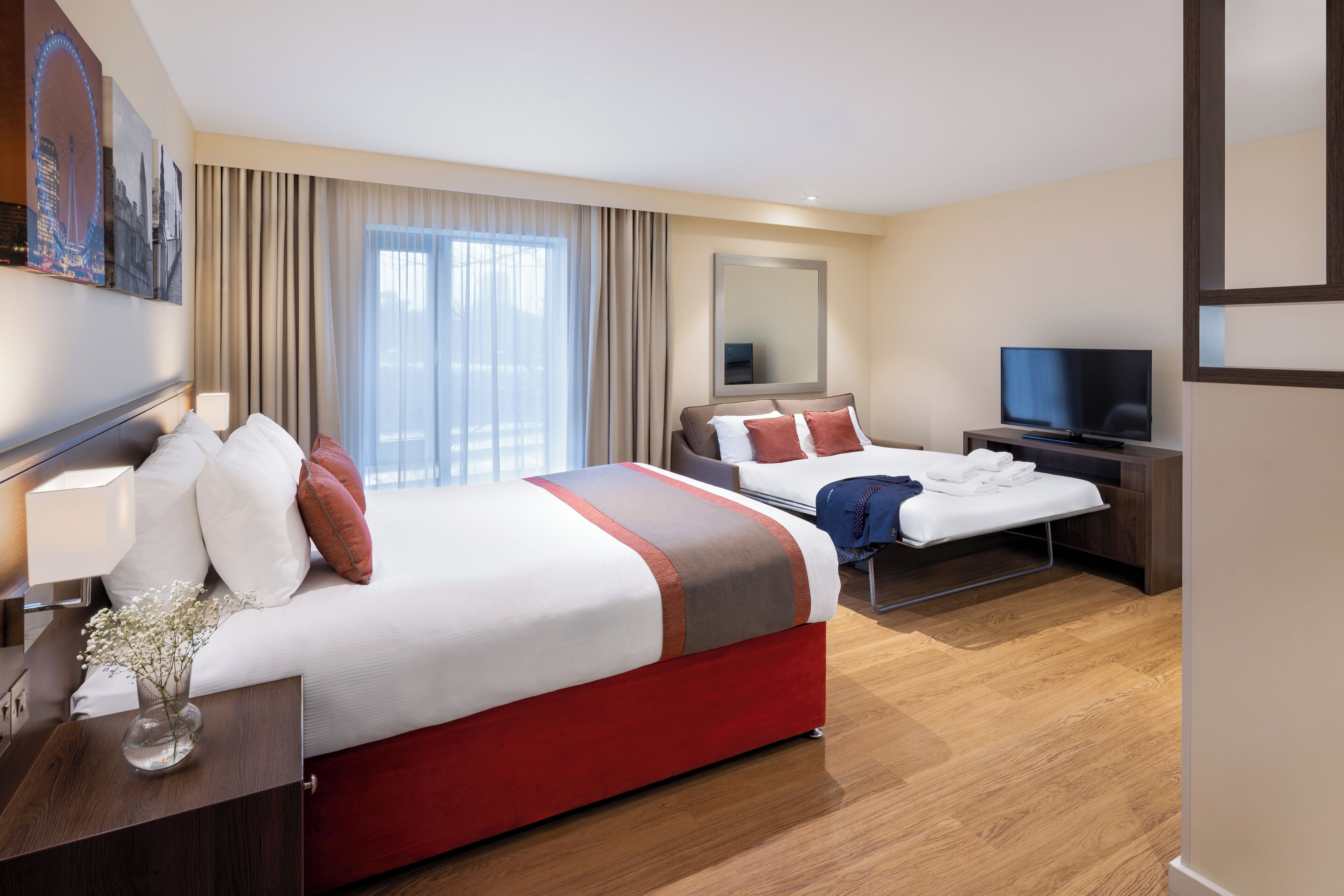 A hotel room with a large bed, a smaller bed, a TV, and a desk with a lamp.