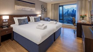 Une chambre d’hôtel moderne, dotée d’un grand lit, d’un bureau et offrant une vue sur le paysage urbain.