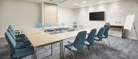 Een vergaderruimte met een lange tafel, blauwe stoelen, een whiteboard en een flatscreen televisie aan een bakstenen muur.