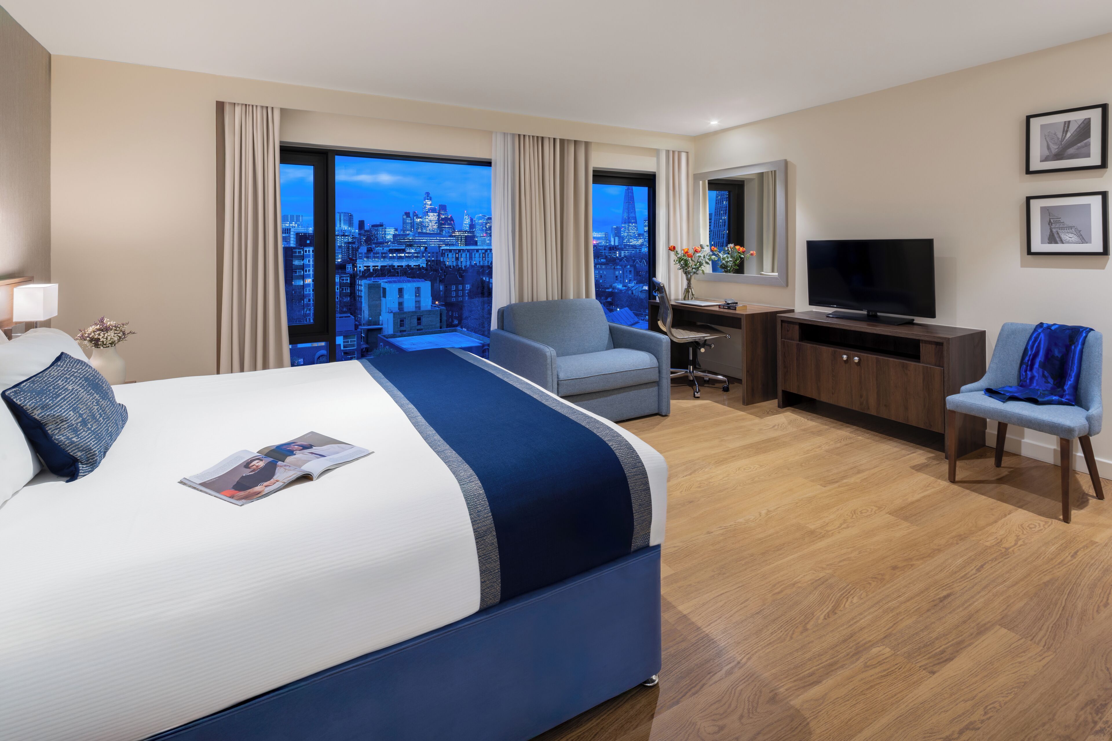 A hotel room with a large bed, a desk, a chair, a television, and a view of the cityscape.