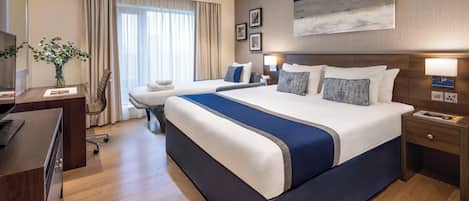 A hotel room with a large bed, a desk, and a chair. There is a window with curtains, a painting on the wall, and a lamp on the nightstand.