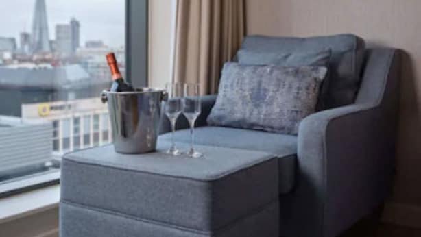 Un salon moderne avec un fauteuil gris, un petit pouf et une vue sur la ville à travers de grandes fenêtres.