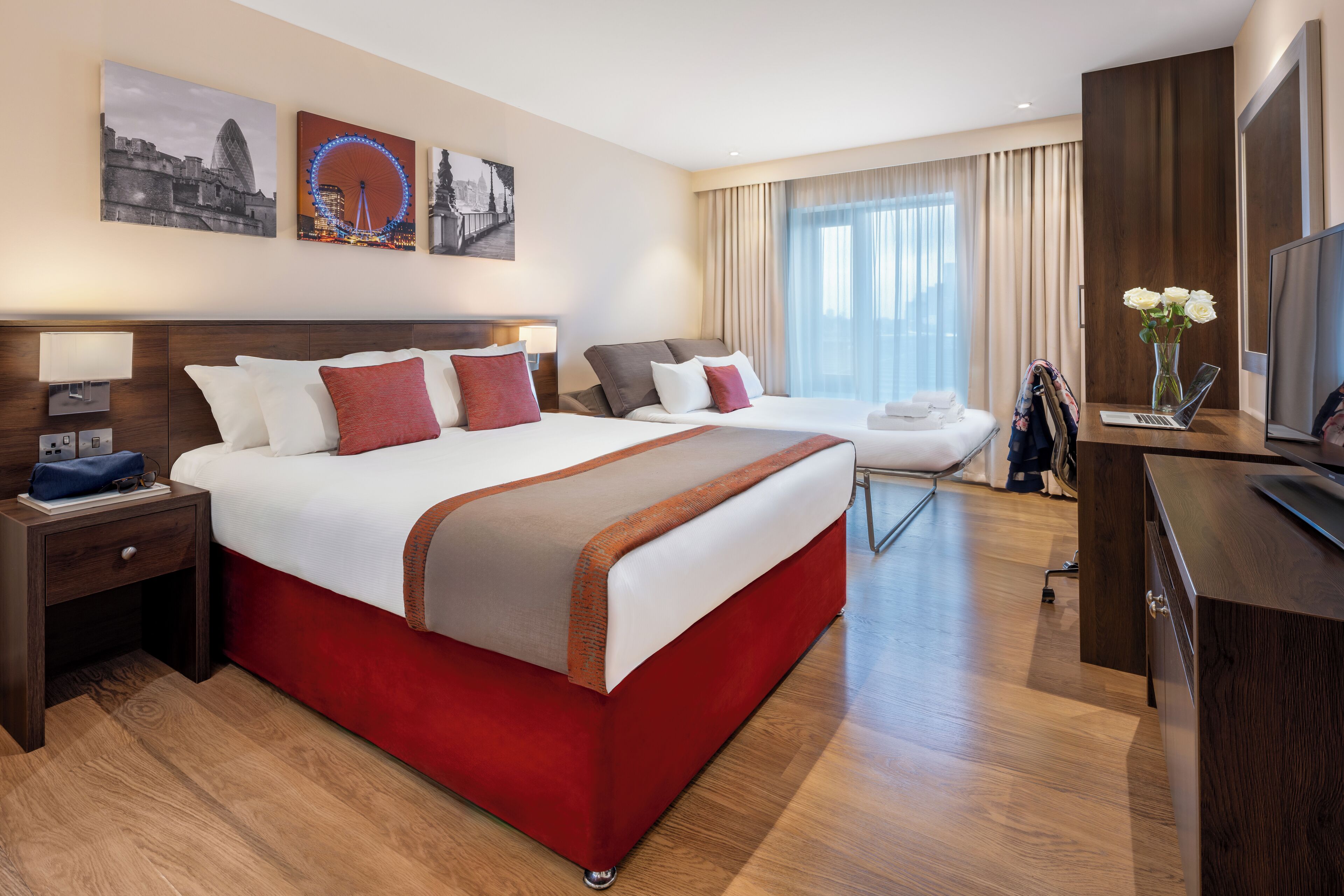 Een hotelkamer met een groot bed, een bureau met een laptop, een televisie en een stoel. Er hangen twee schilderijen aan de muur en staat een vaas met bloemen op het bureau.