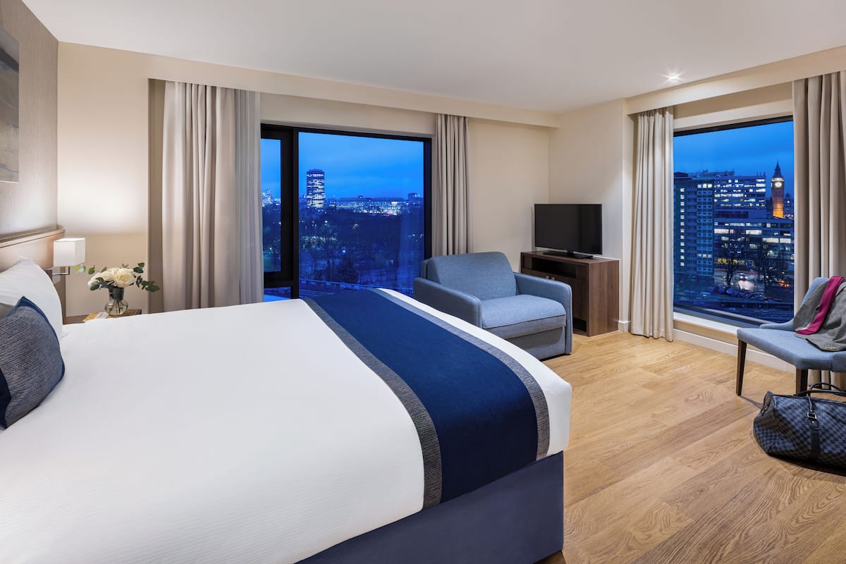 Une chambre d’hôtel avec un grand lit, un coin salon avec un fauteuil, une télévision et une vue sur le paysage urbain au crépuscule.