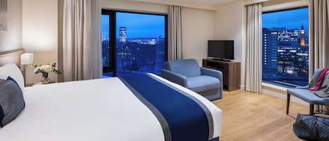 A hotel room with a large bed, a seating area with a chair, a TV, and a view of the cityscape at dusk.