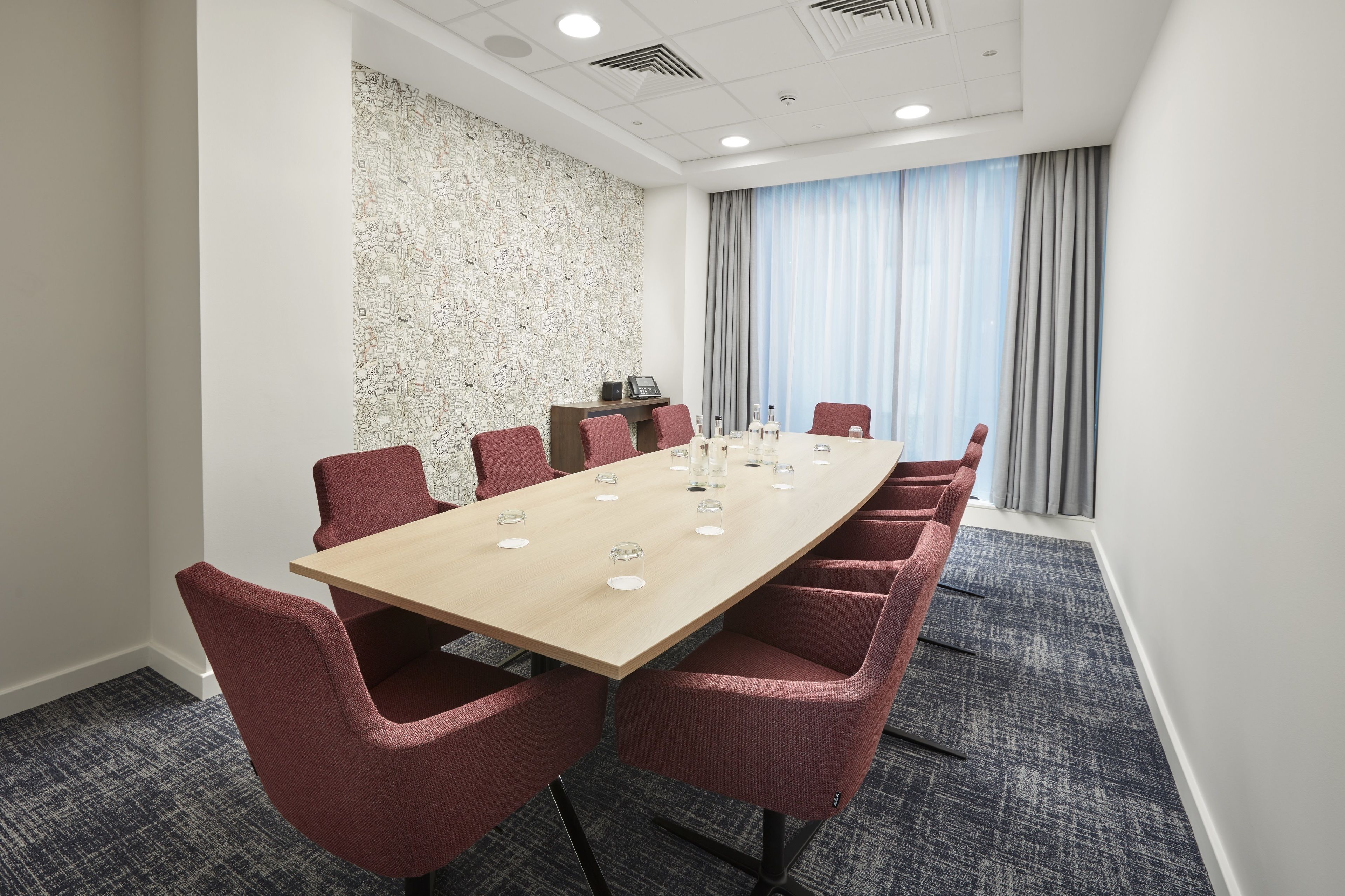 A conference room with a long table, red chairs, a patterned wall, and a window with curtains.