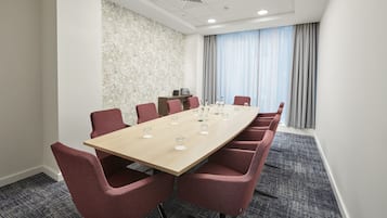 Un salón de conferencias con una larga mesa, sillas rojas, un papel tapiz estampado y una ventana con cortinas.