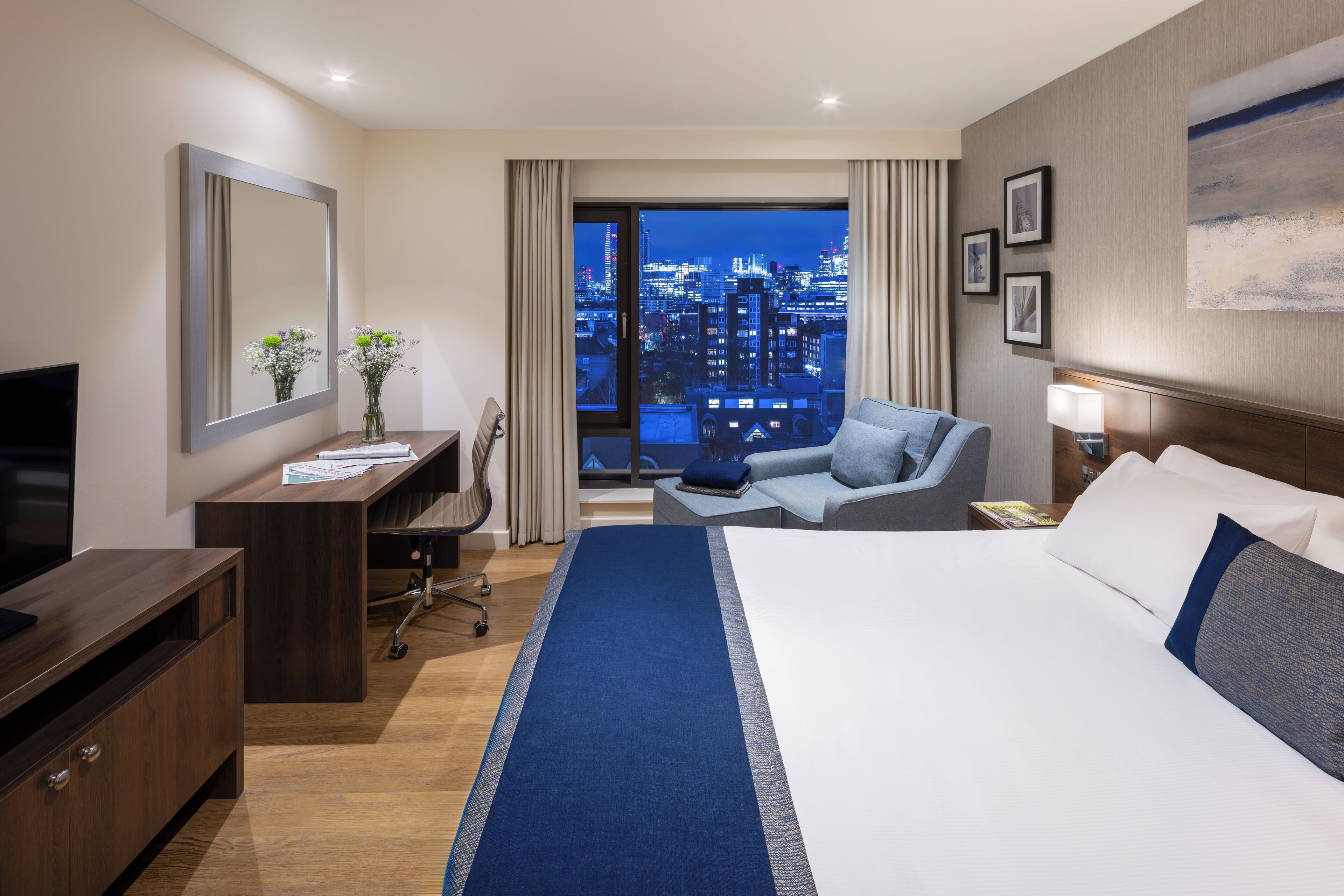 A hotel room with a large bed, a desk with a computer, a chair, a TV, and a view of the city at night.