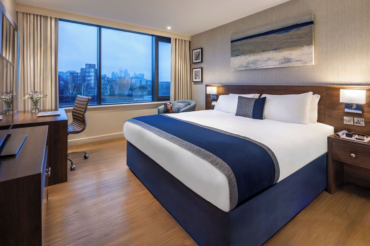 Une chambre d’hôtel avec un grand lit, un bureau avec un ordinateur portable, une chaise et une vue sur la ville grâce à de grandes fenêtres.