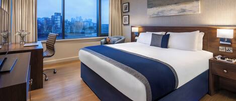 A hotel room with a large bed, a desk with a laptop, a chair, and a view of the city through large windows.