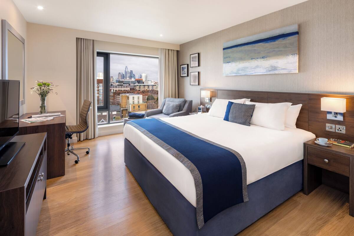 Une chambre d’hôtel avec un grand lit, un bureau avec un ordinateur, une chaise, une télévision et une fenêtre donnant sur la ville.