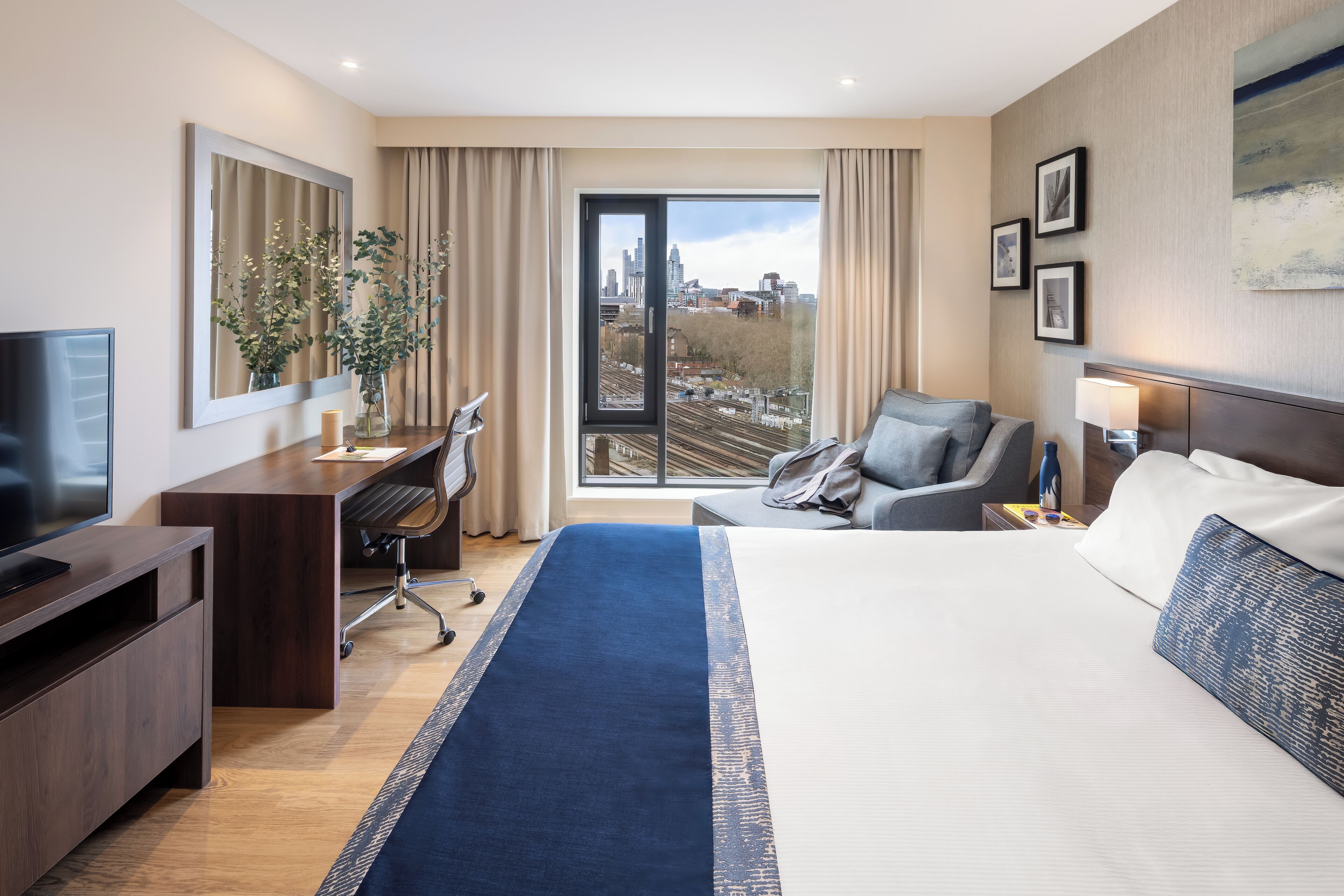 A modern hotel room with a large bed, a desk with a computer, a television, and a view of the city through a large window.