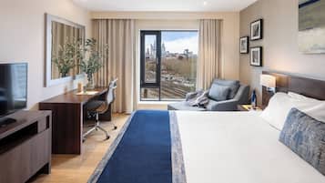 A modern hotel room with a large bed, a desk with a computer, a television, and a view of the city through a large window.