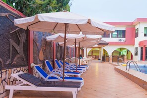 A poolside area with lounge chairs and umbrellas, a pool, and a building with arches and a stone wall. - Kesdem Hotel (Tema)