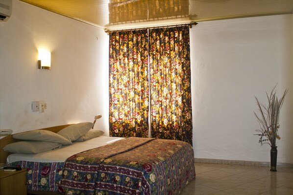 Un dormitorio con cama, una mesita de noche, una lámpara de pared y un ventanal con cortinas florales.