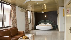 Una habitación de hotel con cama, sofá, una mesita, ventilador de techo y ventana con cortinas.
