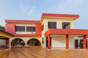 A modern two-story building with a red and beige facade, featuring an open courtyard with tiled flooring and outdoor seating. - Kesdem Hotel (Tema)