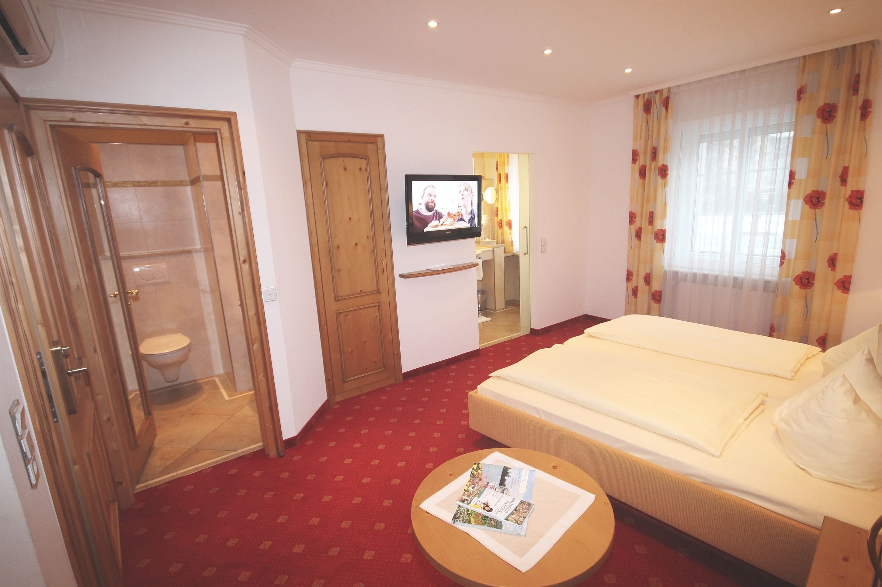 A hotel room with a bed, a small table with magazines, a television on the wall, and a bathroom with a toilet and shower.