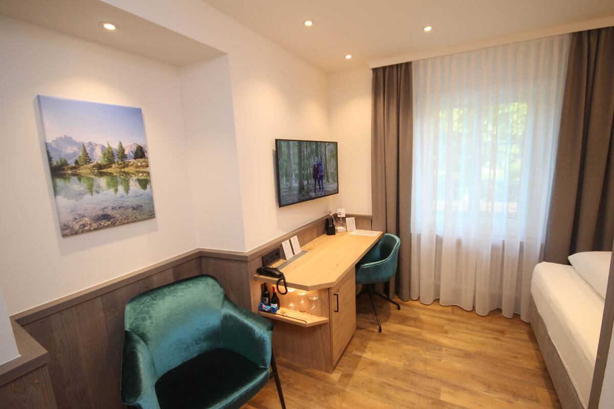 Habitación de hotel con un escritorio de madera, una silla, una cama, un televisor montado en la pared y un cuadro de un paisaje montañoso.