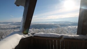 Una terraza de madera cubierta de nieve, con vista a un paisaje nevado y edificios lejanos bajo un cielo despejado.