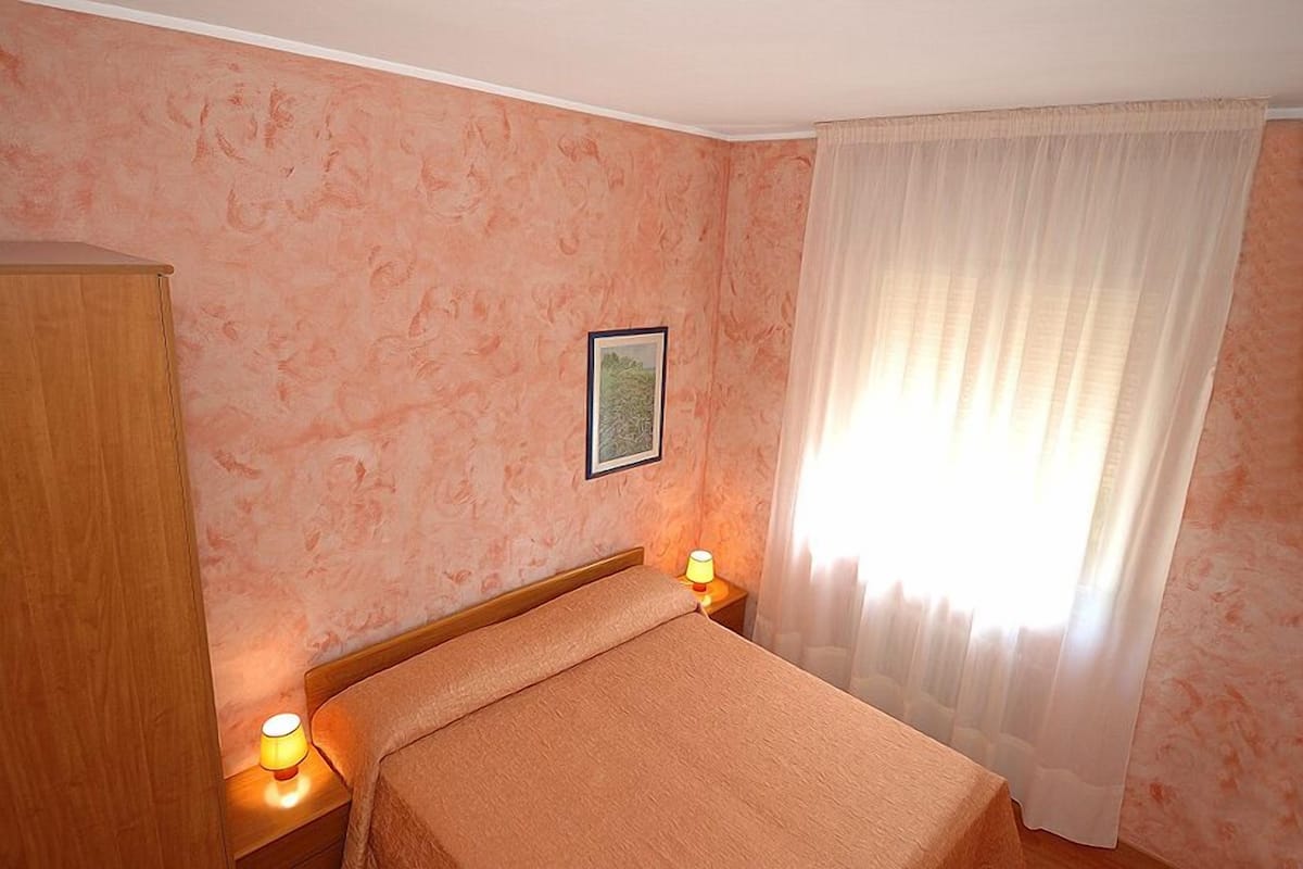 Una habitación de hotel con cama, mesita de noche con lámpara, armario y ventana con cortinas traslúcidas.