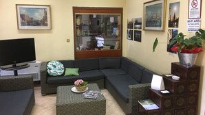 A living room with a sectional sofa, a television, wicker furniture, and a view of the city through a glass door. - Primavera Hotel (Mestre)
