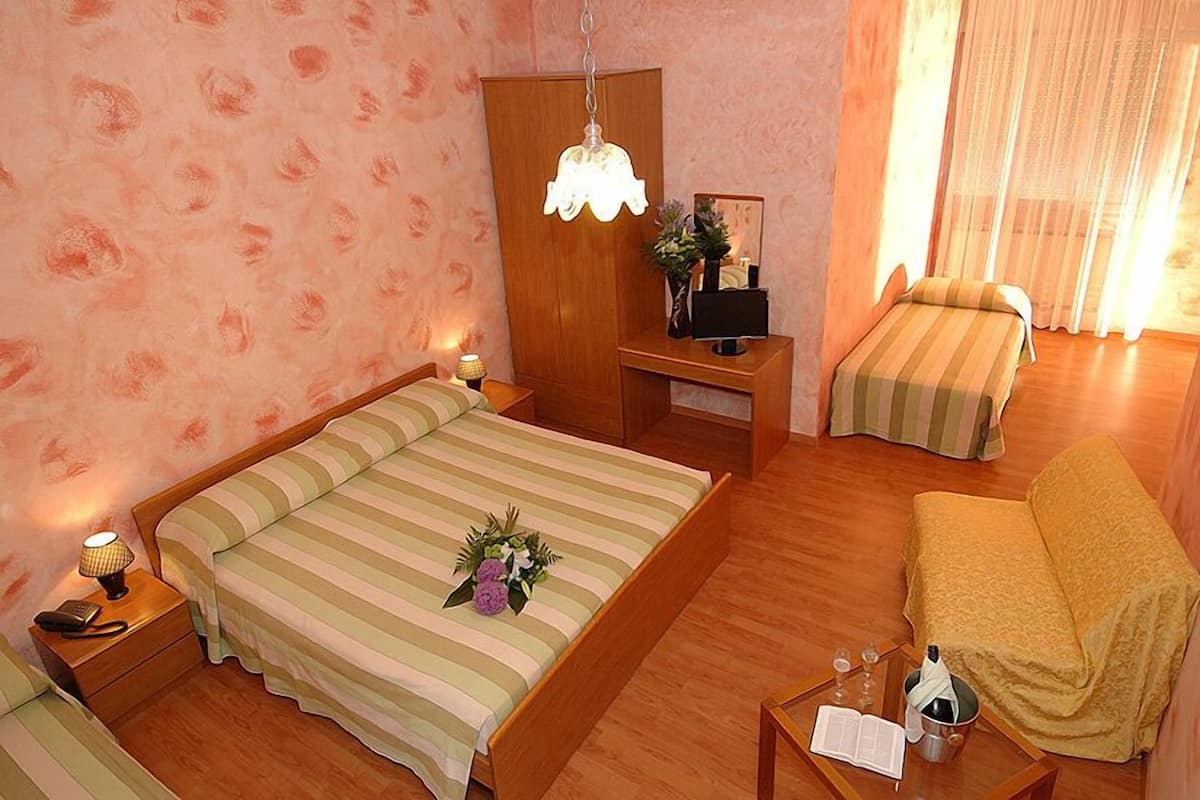 Una habitación de hotel con una cama grande, otra más pequeña, un sofá, una mesita de noche con lámpara, una mesita auxiliar y una pequeña mesa con una botella y vasos.