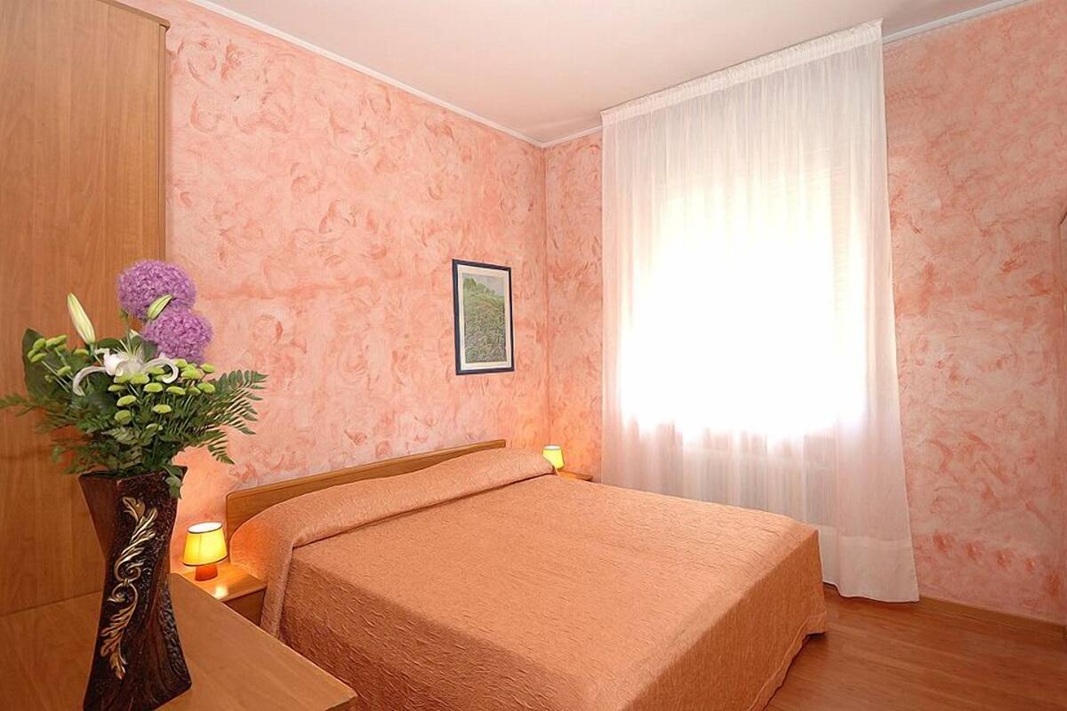 Habitación de hotel con una cama, una mesita de noche con lámpara, un jarr ón con flores y una ventana con cortinas.