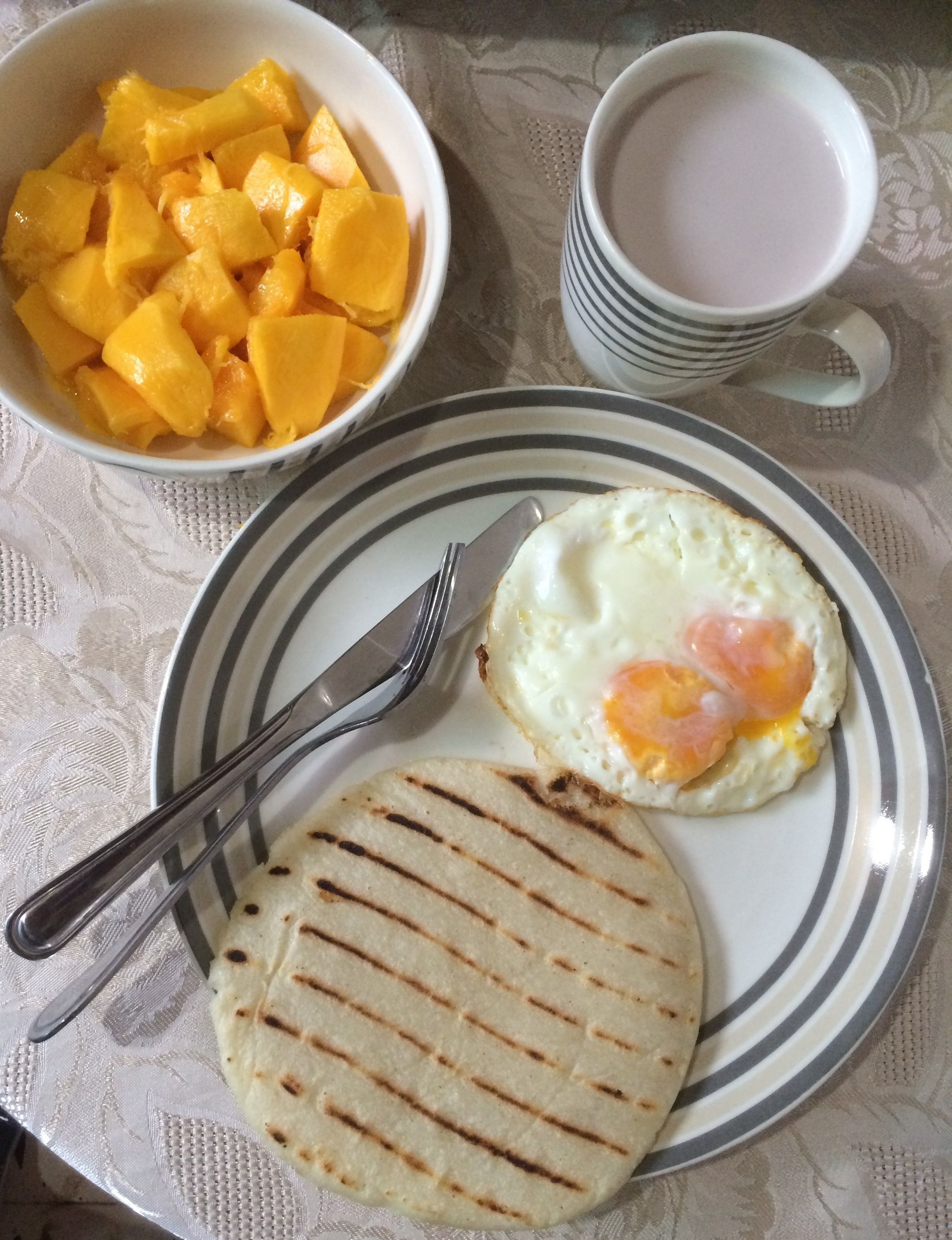 Un plato con un pan plano a la parrilla, un huevo frito y un tazón de mango en cubos sobre una mesa con mantel de encaje.