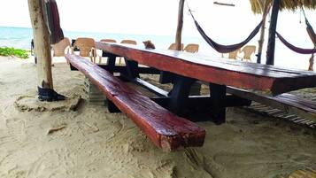 Un área con bancos de madera frente al mar, cubierta por un techo de paja y rodeada de sillas de plástico.