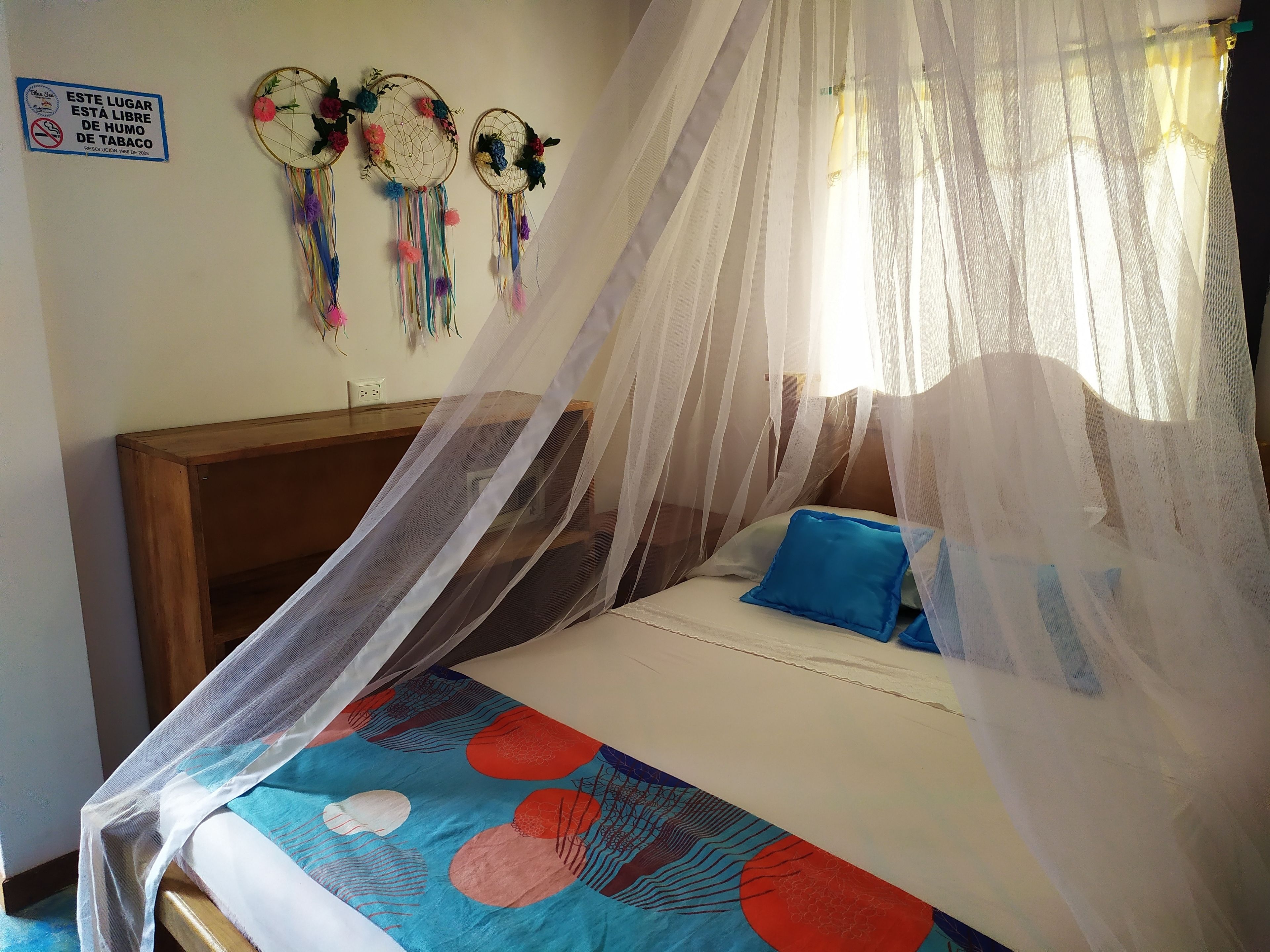 Una cama con dosel, una pared decorada con atrapasueños y un cartel de prohibido fumar.
