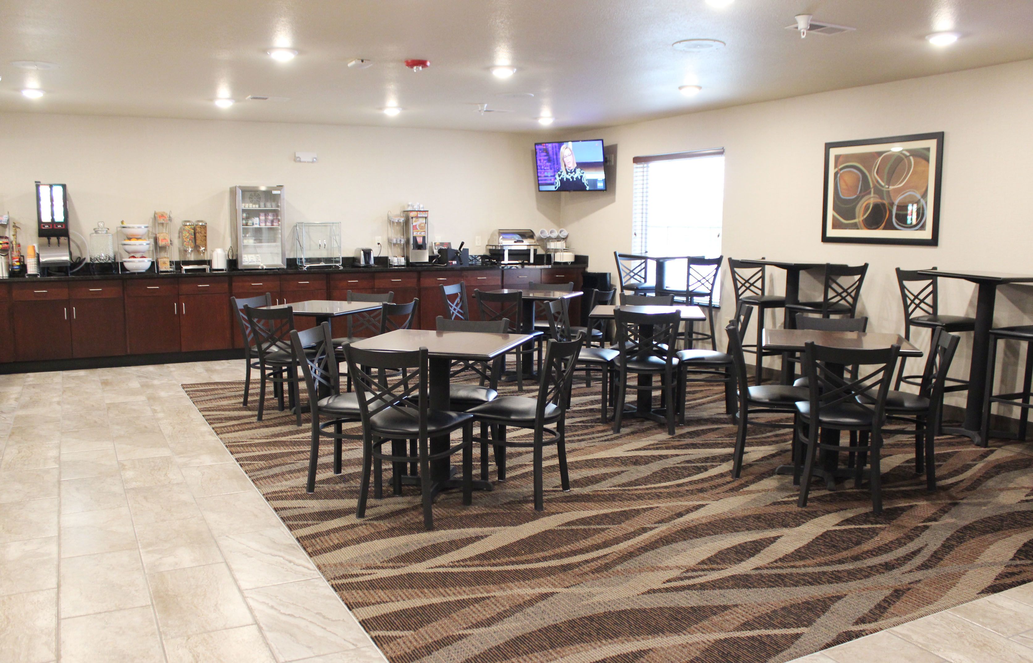 A hotel dining area with multiple tables and chairs, a kitchen counter, and a television mounted on the wall.
