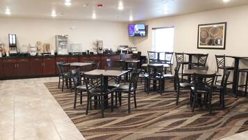 A hotel dining area with multiple tables and chairs, a kitchen counter, and a television mounted on the wall.