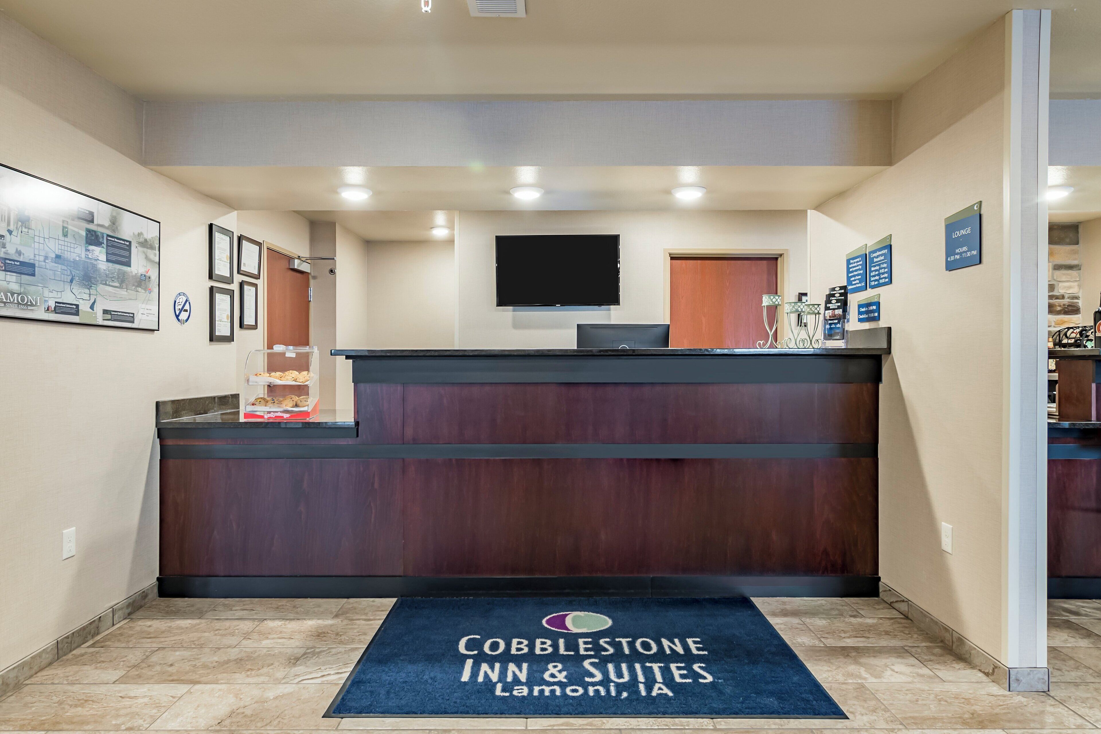 A reception area with a wooden reception desk, a flat-screen TV mounted on the wall, and a blue mat with the hotel name.