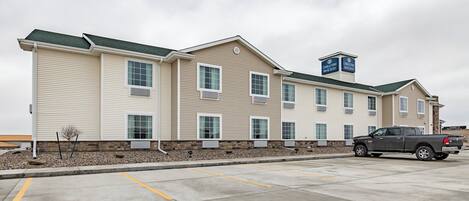 A multi-story hotel with a beige exterior and green roof. There is a parking lot with a few vehicles and a sign indicating hotel services.