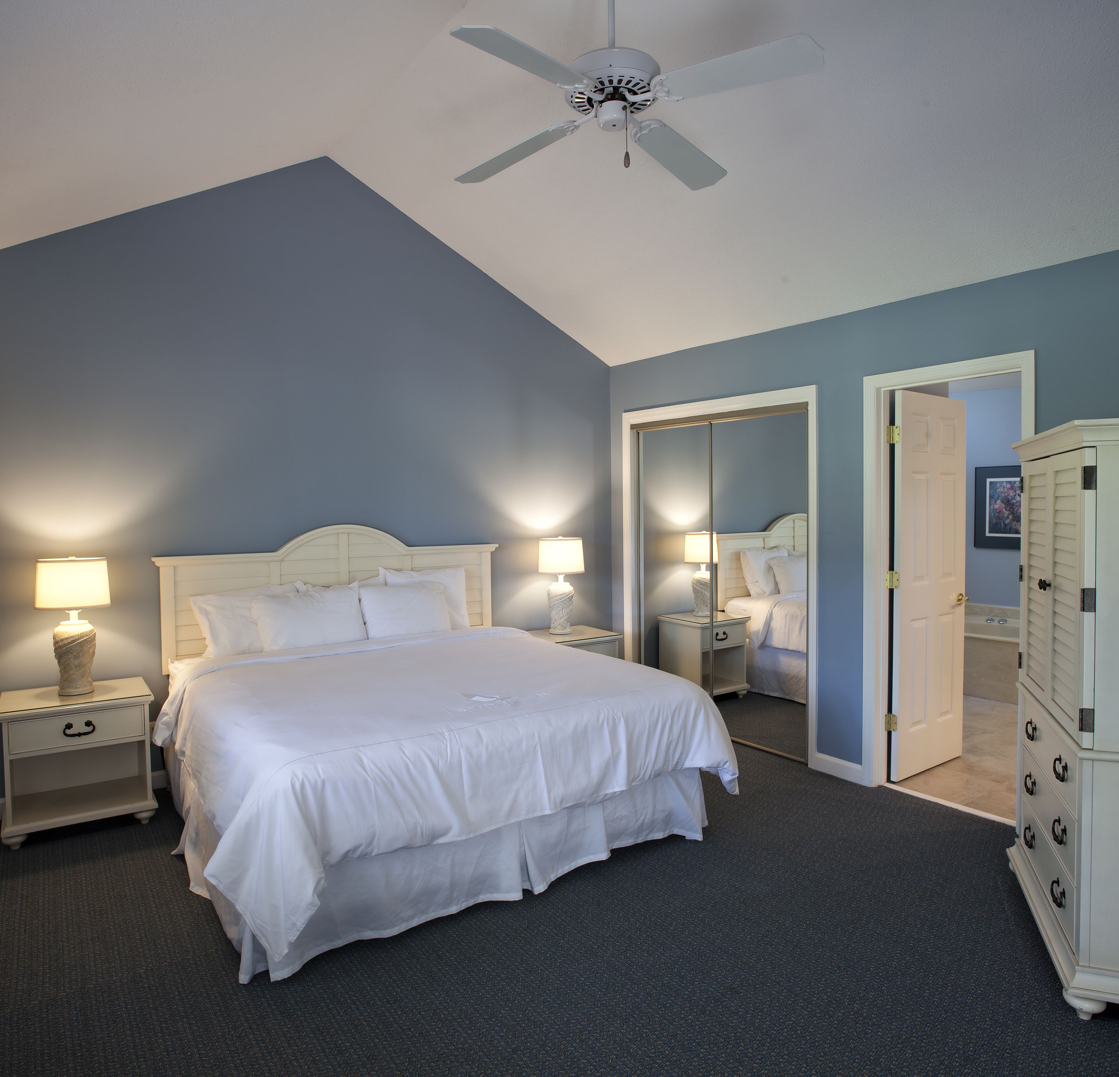 A bedroom with a bed, two bedside tables with lamps, a ceiling fan, a mirror, and a walk-in closet.