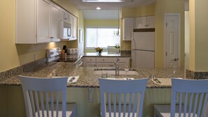 A kitchen with white cabinets, granite countertops, and a central island with two white chairs.