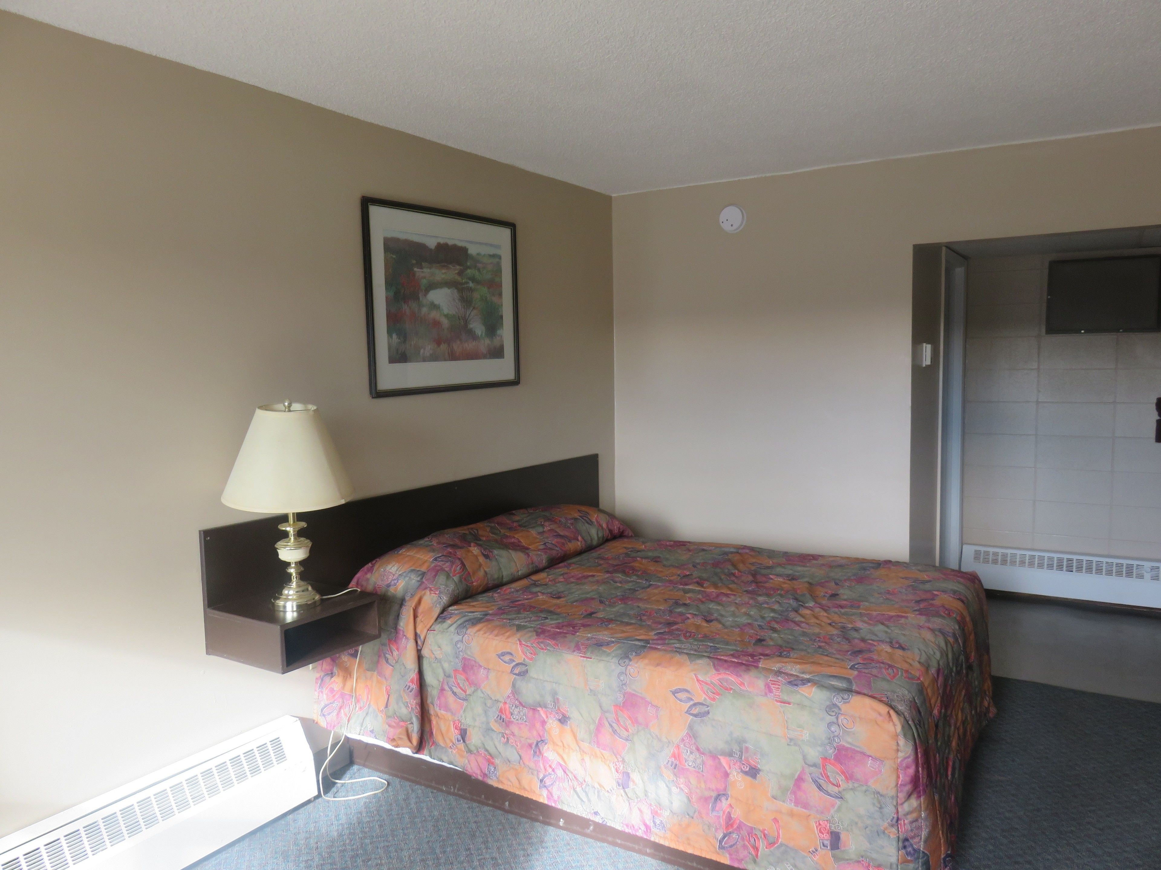 Una camera d'albergo con un letto, un comodino con una lampada, un quadro appeso al muro e una porta sulla destra.