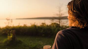 Une personne tenant une tasse, debout à l’extérieur, avec une étendue d’eau et de la verdure en arrière-plan.