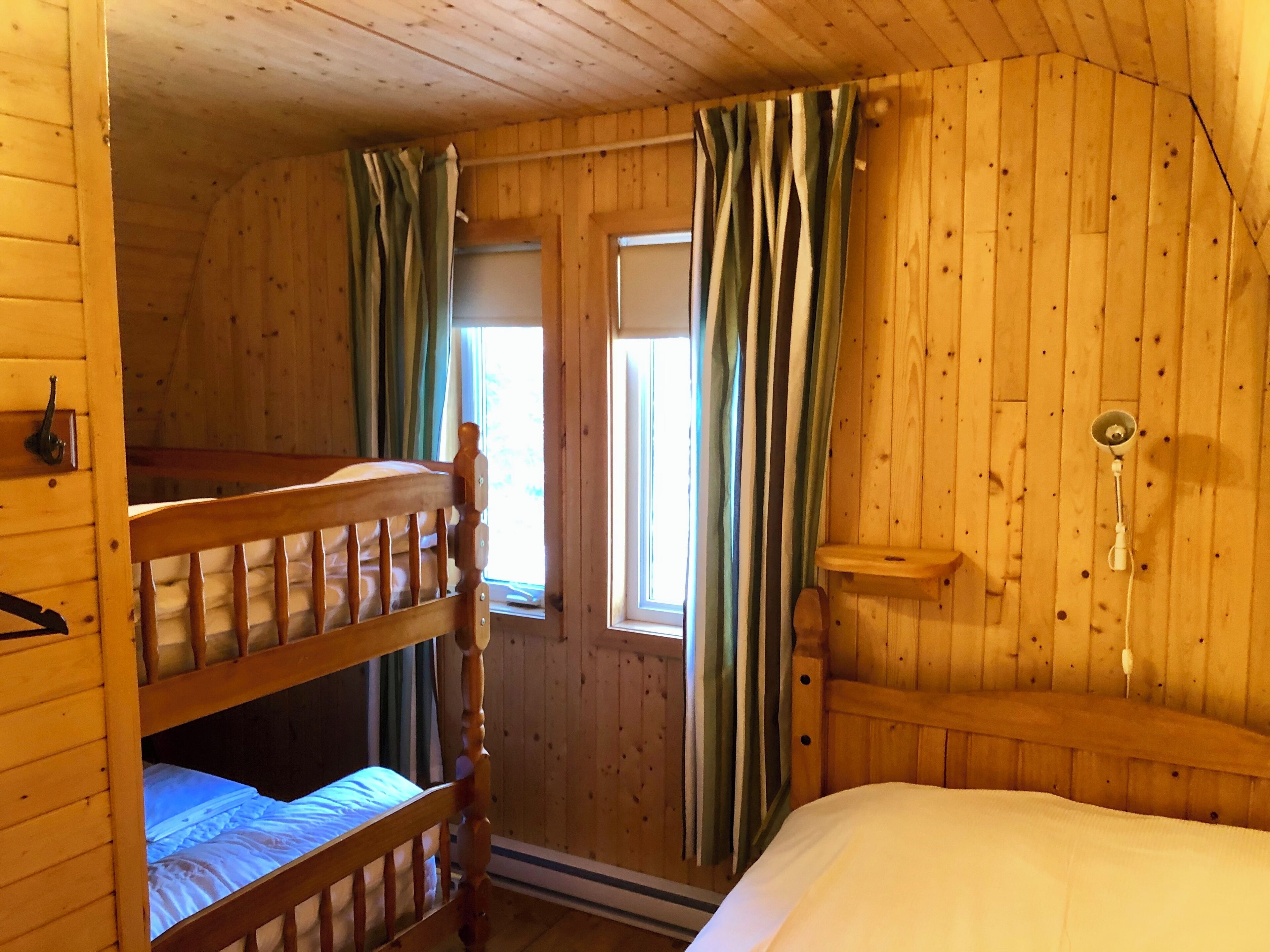 Une pièce de cabane en bois avec des lits superposés, une fenêtre avec des rideaux et un luminaire fixé au mur.