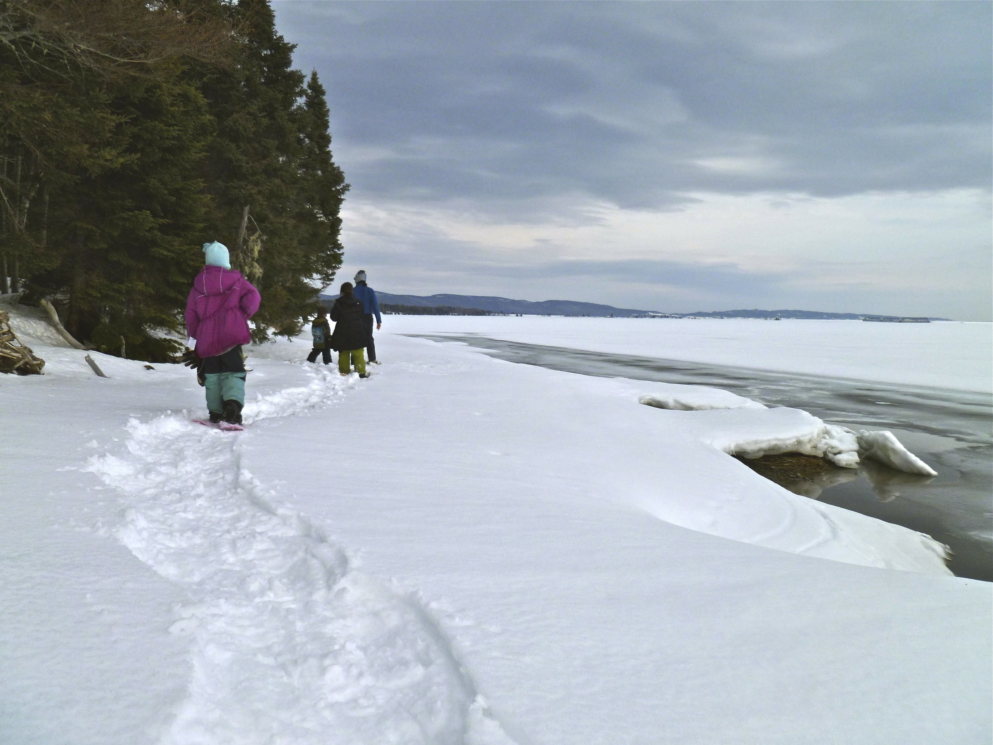 A snow-covered path beside a frozen body of water with trees on one side.