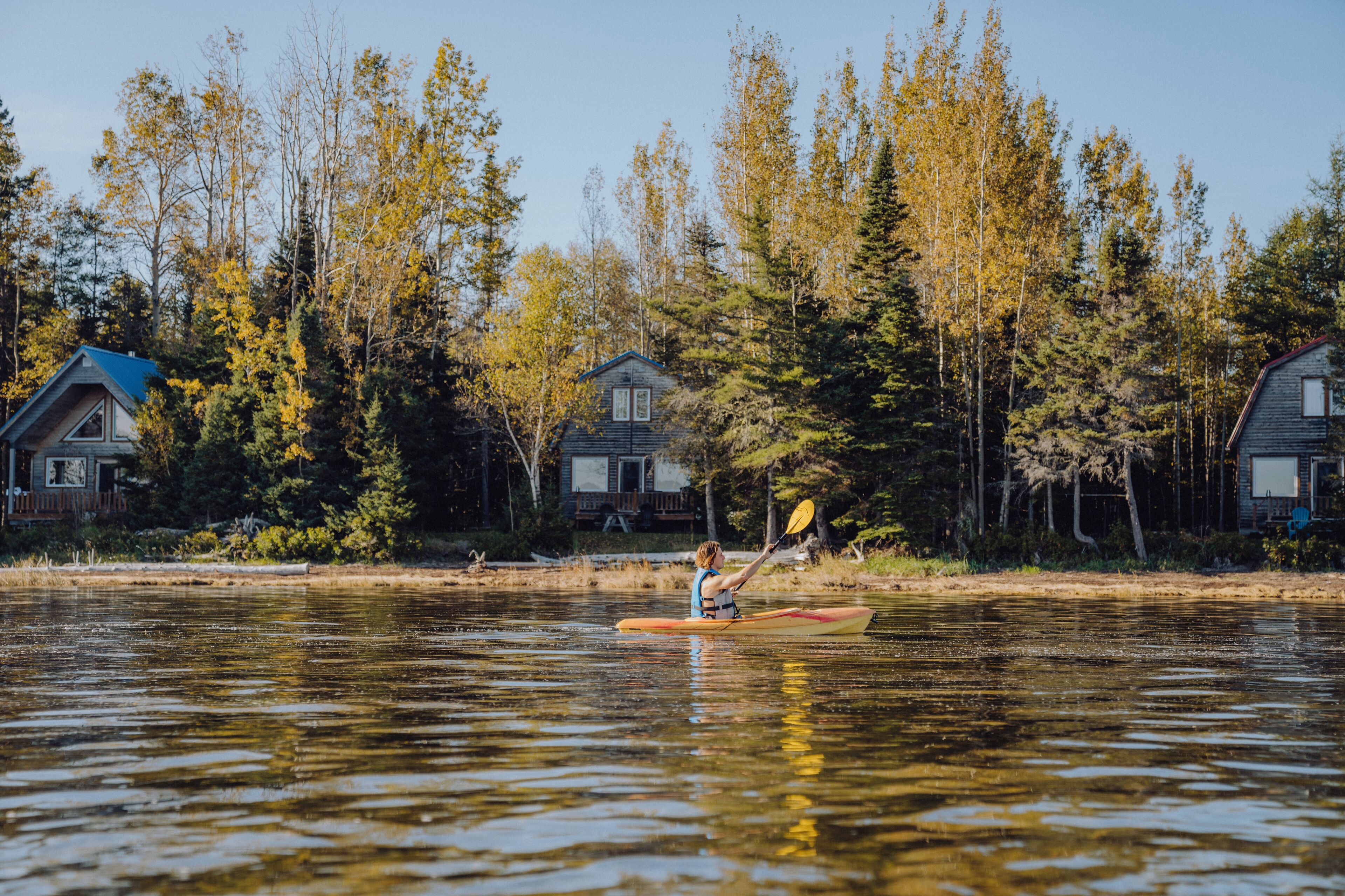 A person kayaking on a calm lake with houses and trees in the background.