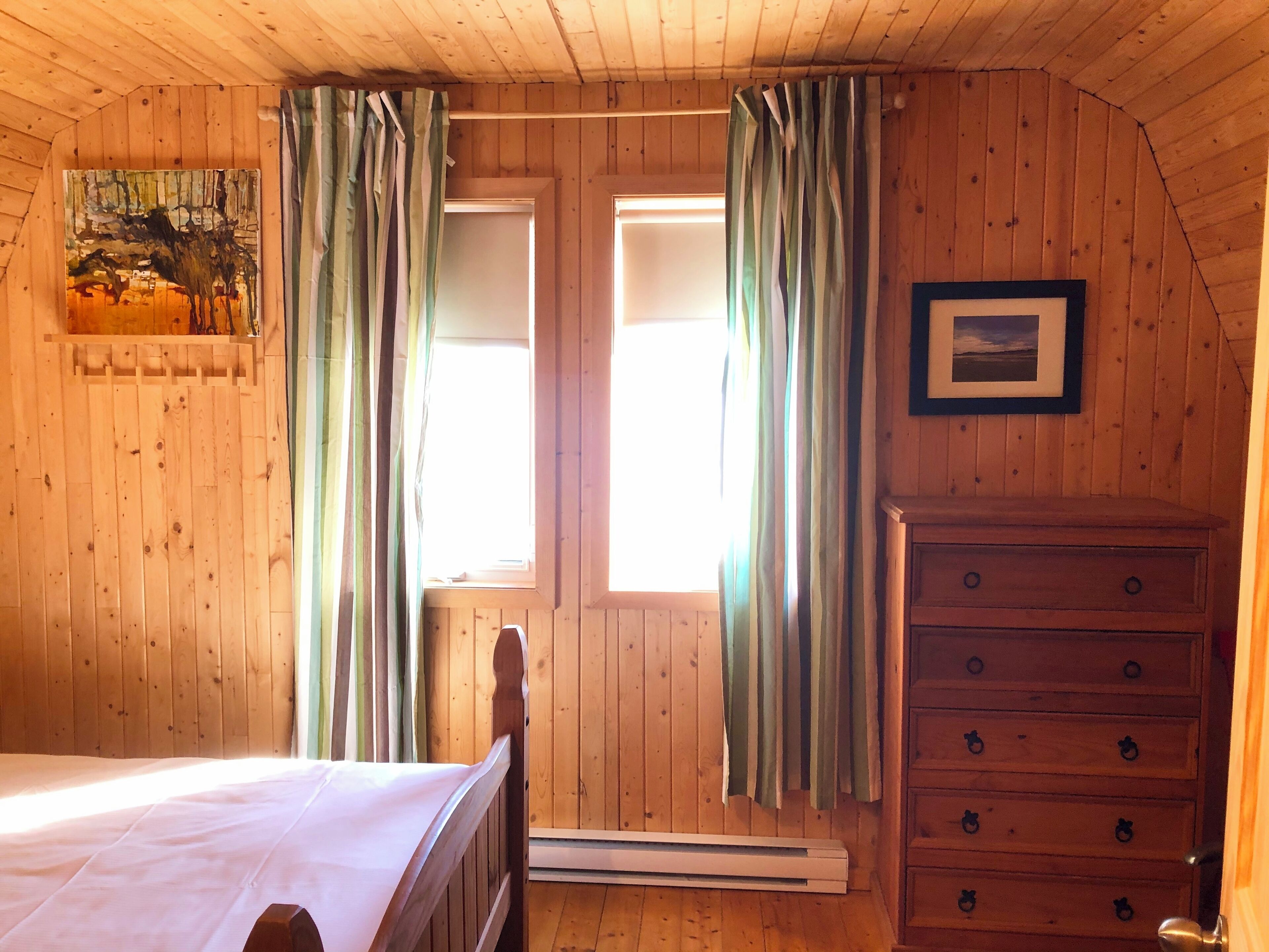 A wooden room with a bed, a dresser, and two windows with curtains.